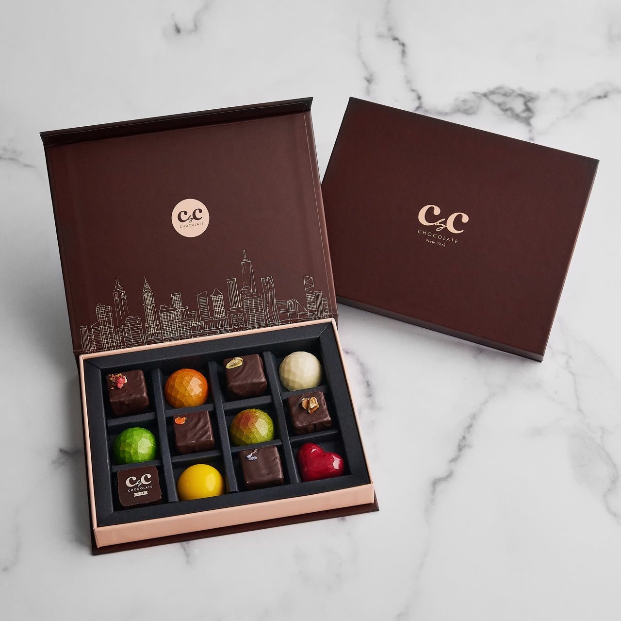 12-piece CbyC Chocolate Bonbon Box with assorted colorful chocolates neatly arranged in a brown box featuring a gold city skyline design.