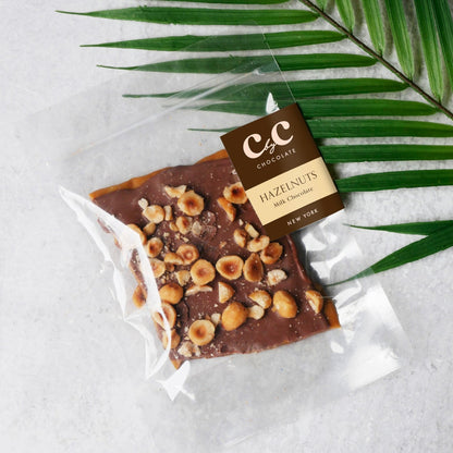 C by C New York Hazelnuts Milk Chocolate Toffee — a piece of milk chocolate bark topped with roasted hazelnuts, elegantly packaged in clear wrapping beside a green palm leaf on a light stone surface.