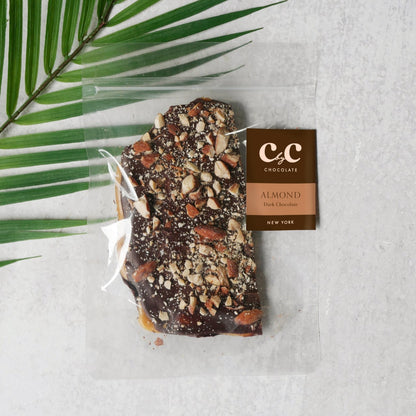 C by C New York Almond Dark Chocolate Toffee — a rich dark chocolate bark topped with roasted almond pieces, elegantly packaged in clear wrapping beside a green palm leaf on a light stone surface.