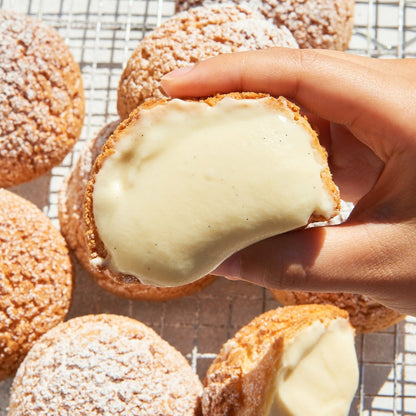 Hand holding a cream puff broken open to reveal smooth vanilla cream filling, surrounded by baked cream puffs.