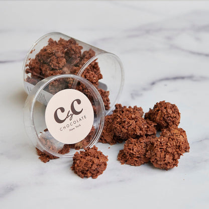 A clear plastic container with chocolate covered crunchy treats spilling out onto a marble surface, with the brand label 'C by C New York' visible on the container.