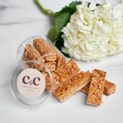 C by C New York Florentines displayed on a marble surface, with golden almond toffee layers spilling from a clear jar beside a white hydrangea blossom.