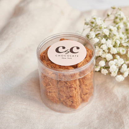 Transparent jar of C by C New York Florentines placed beside white flowers on a soft beige fabric background.