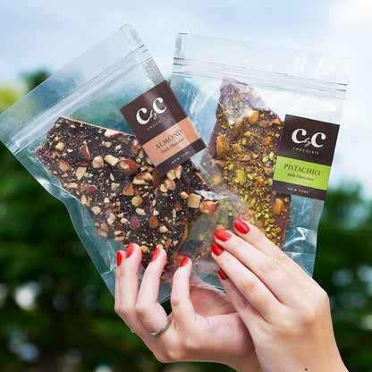 Hands holding two C by C New York chocolate toffee packs — Almond Dark Chocolate and Pistachio Milk Chocolate — against a softly blurred green outdoor background, showcasing their elegant packaging and nut toppings.