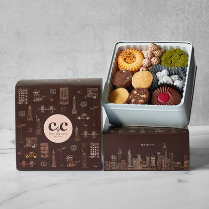 C by C C New York Assorted Cookie Tin featuring a colorful variety of handmade cookies, elegantly presented in a light gray tin beside a brown New York–themed gift box with gold illustrations.
