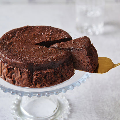 A flourless chocolate cake on a glass pedestal with a slice being served using a gold cake server.