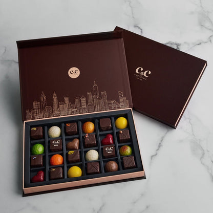 24-piece CbyC Chocolate Bonbon Box with vibrant assorted bonbons, opened on a marble surface, with hands selecting a piece.