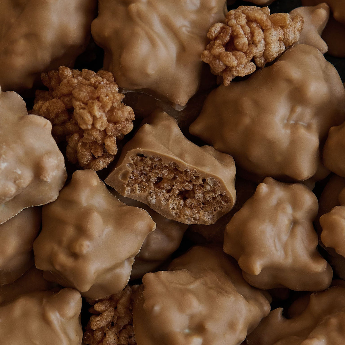 A close-up pile of chocolate-covered cereal clusters, with one piece broken open to show the textured rice cereal interior.