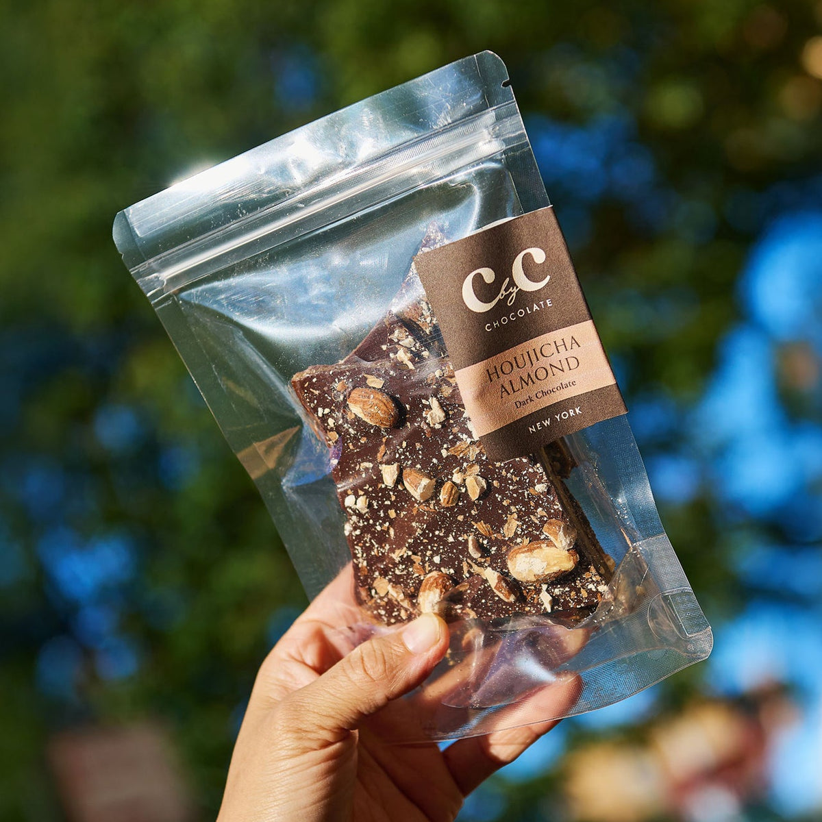A hand holds a clear stand-up pouch of chocolate almond bark outdoors, with blurred green foliage and blue sky in the background. The pouch has a brown label reading 'CC Chocolate Company' and 'Almondia Almond Butter'.