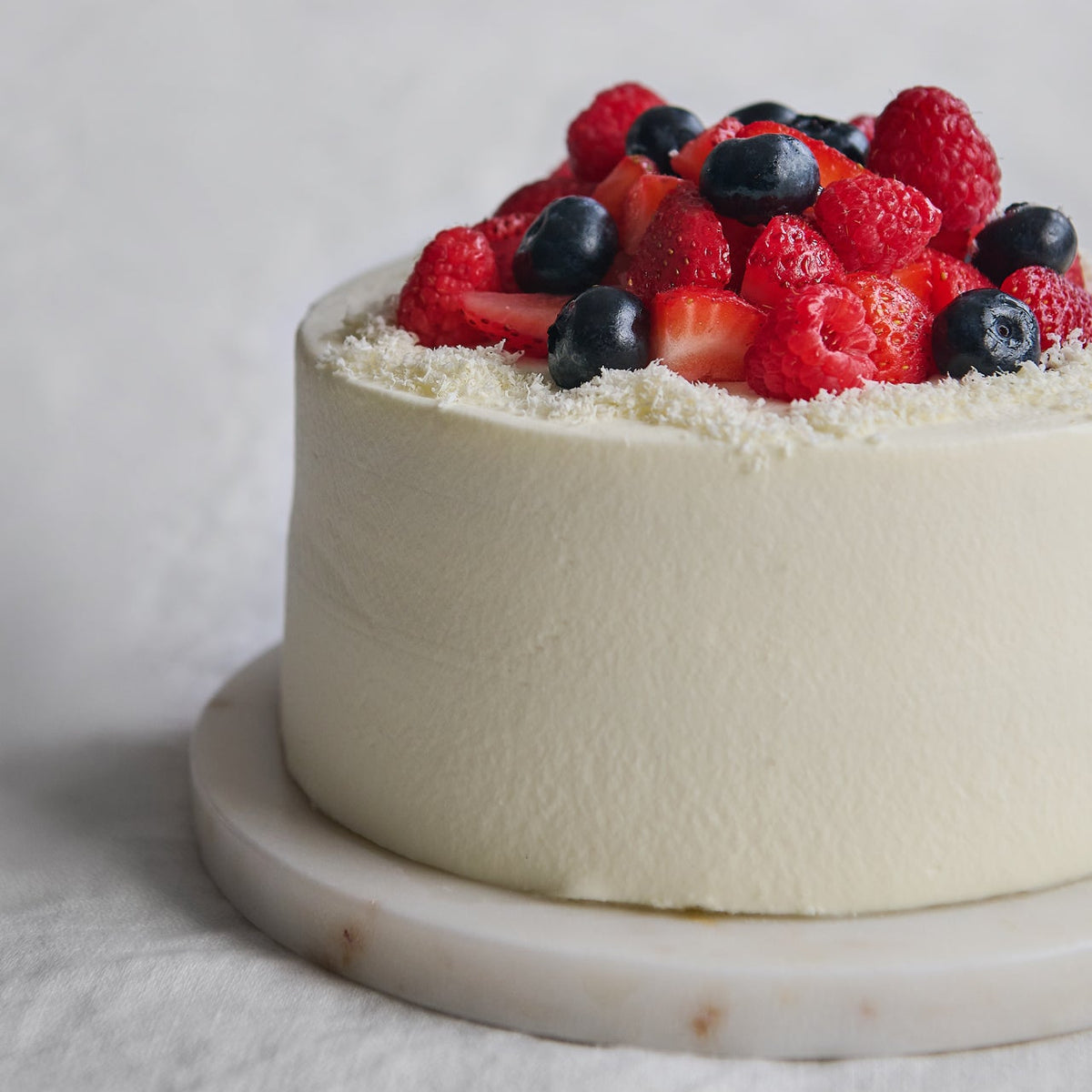 A round white frosted cake on a marble stand, topped with fresh red strawberries, raspberries, and blueberries, photographed against a soft light gray background.
